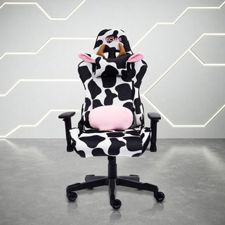 Front view of the techni sport cow gaming chair in black and white in a white room with grey floor. This cow print gaming chair features a black base. The cow chair headrest pillow has light brown horns and ears lined with fluffy pink material and the lumbar pillow has matching fluffy pink to compliment the design. This is definitely one of the cutest Techni Sport Gaming chair options and a great option for cute gaming setups or home office setups. 