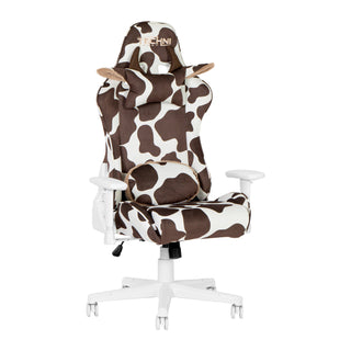 The Techni Sport cow gaming chair in brown shown against a white background at an angle. The brown cow gaming chair has the lumbar pillow flipped around to show that it is reversible with brown and white print on one side and fluffy beige material on the back. The brown cow gaming chair also features a white base, wheels and hardware. The chocolate cow gaming chair also has a headrest with brown horns and cute ears. The materials used are velvet for the ultimate softness. 