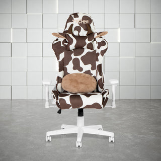 Front view of the cow gaming chair in brown in a white room with grey floor. This cow gaming chair features a white base, with white and brown cow print. The cow chair headrest pillow has brown horns and ears lined with fluffy beige material and the lumbar pillow has matching fluffy beige to compliment the design. This is definitely one of the cutest Techni Sport Gaming chair options and a great option for kawaii gaming setups or as a cute wfh chair. 