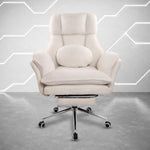 A cream-colored high-back lounge chair with a matching round cushion and footrest, set on a chrome rolling base against a modern gray wall with geometric light accents. 