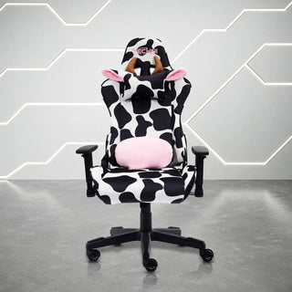 Front view of the techni sport cow gaming chair in black and white in a white room with grey floor. This cow gaming chair features a black base, with white and black cow print. The cow chair headrest pillow has light brown horns and ears lined with fluffy pink material and the lumbar pillow has matching fluffy pink to compliment the design. This is definitely one of the cutest Techni Sport Gaming chair options and a great option for kawaii gaming setups or as a cute home office chair. 