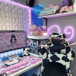 Cute gaming setup with cow print chair, pink and white decor, and computer equipment. 